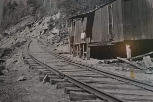 A train tracking heading up a mountain beside a small lumberman's home with a toddler standing in the doorway 