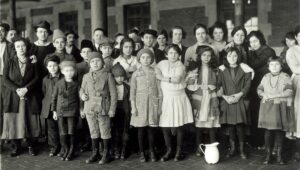 A group of about 40 immigrant adults and children at Ellis Island in 1908. None of them are smiling, and one of the girls on the front row is clutching a doll. 
