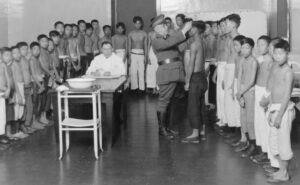 A group of about 40 Asian boys, all with their shirts off, is inpected by a uniformed doctor at Angel Island Immigration Station in San Francisco Bay. None of the boys are smiling.