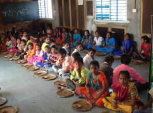 A group of Indian students wearing neat, colorful clothing sitting on the floor of their school at lunchtime. 