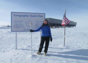 Scientist Katherine Joy stands beside a sign that says "Geographic South Pole."