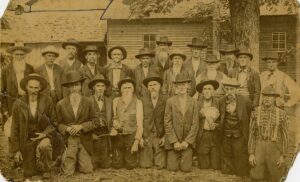 An old photo that shows about 20 older men posing in front of the camera in 1905. All are wearing suits and hats.
