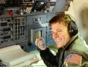 A man smiles while sitting at the navigator seat of a Navy P3 Orion. He has headphones on, and the instruments in front of him look like they are from the 1960s. 