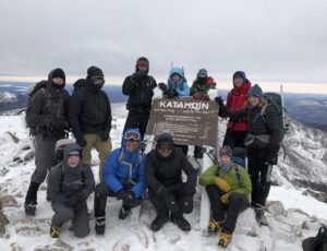 A group of about a dozen hikers bundled up in winter clothing and gather around a sign that says that they are at the top of Mount Katahdin, in Maine. 