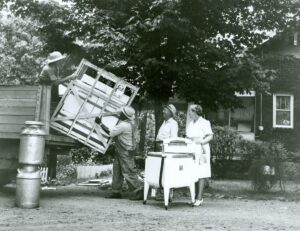 An older couple stand in front of their home while workers unload appliances at their home in Tennessee in the 1940s 
