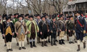 A group of Colonial reenactors in period costume and carrying guns over their shoulders, line up to march. 