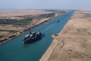 A massive aircraft carrier goes through the Suez Canal. The water is blue. The desert on both sides of the canal is brown. 
