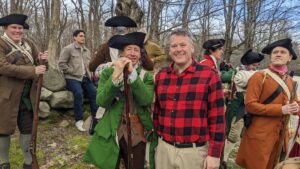 A smiling man in a red flannel shirt poses and smiles with colonial reenactors in Concord, Massachusetts. 